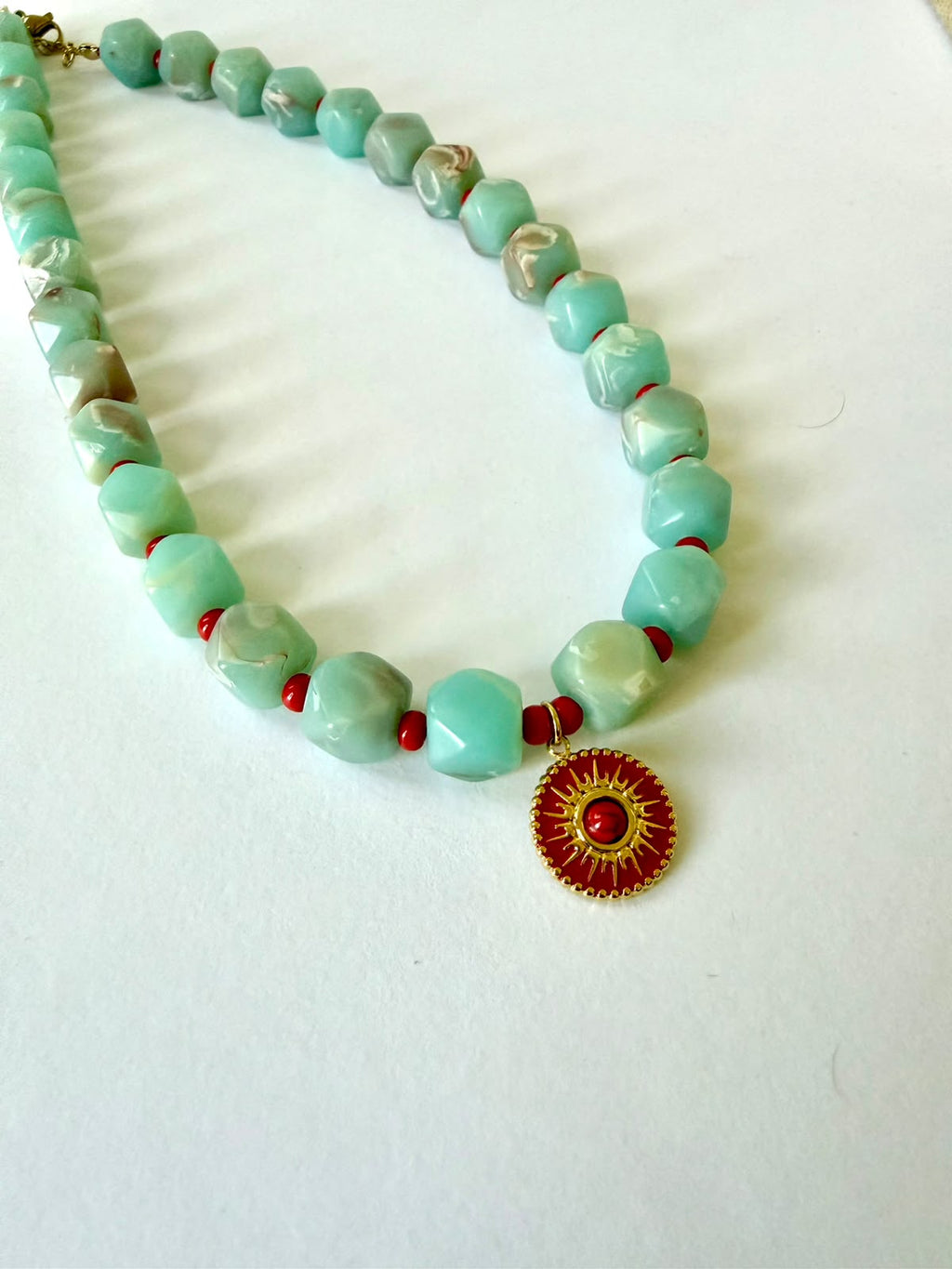 Close-up of faceted aqua acrylic beads with marbled details, small red beads, and a round red pendant with a gold-tone sun design.