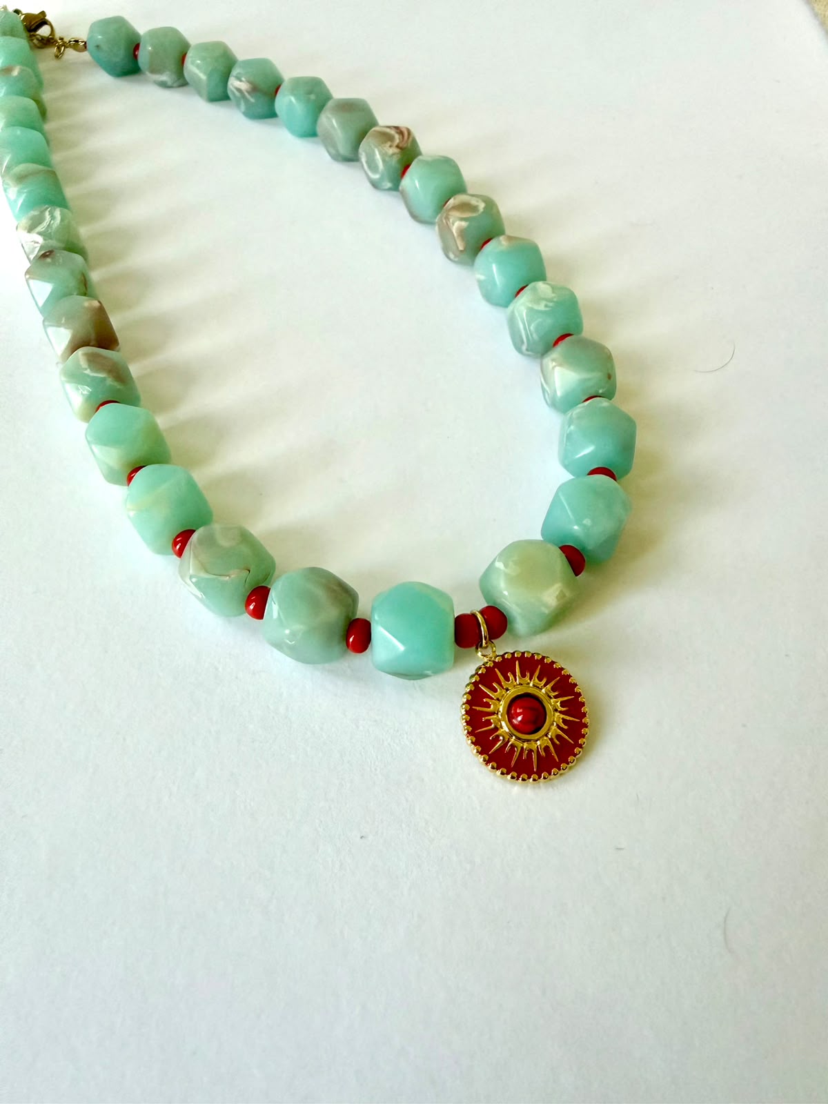 Close-up of faceted aqua acrylic beads with marbled details, small red beads, and a round red pendant with a gold-tone sun design.