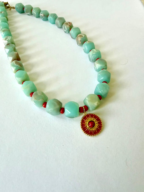 Close-up of faceted aqua acrylic beads with marbled details, small red beads, and a round red pendant with a gold-tone sun design.