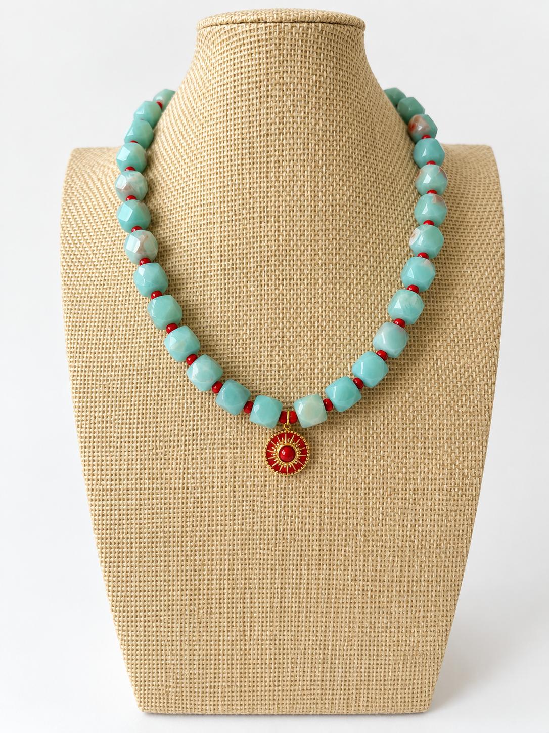 Aqua acrylic beaded necklace with small red beads and a red and gold-tone sun pendant on a beige jewelry bust.