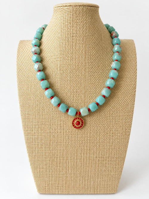 Aqua acrylic beaded necklace with small red beads and a red and gold-tone sun pendant on a beige jewelry bust.