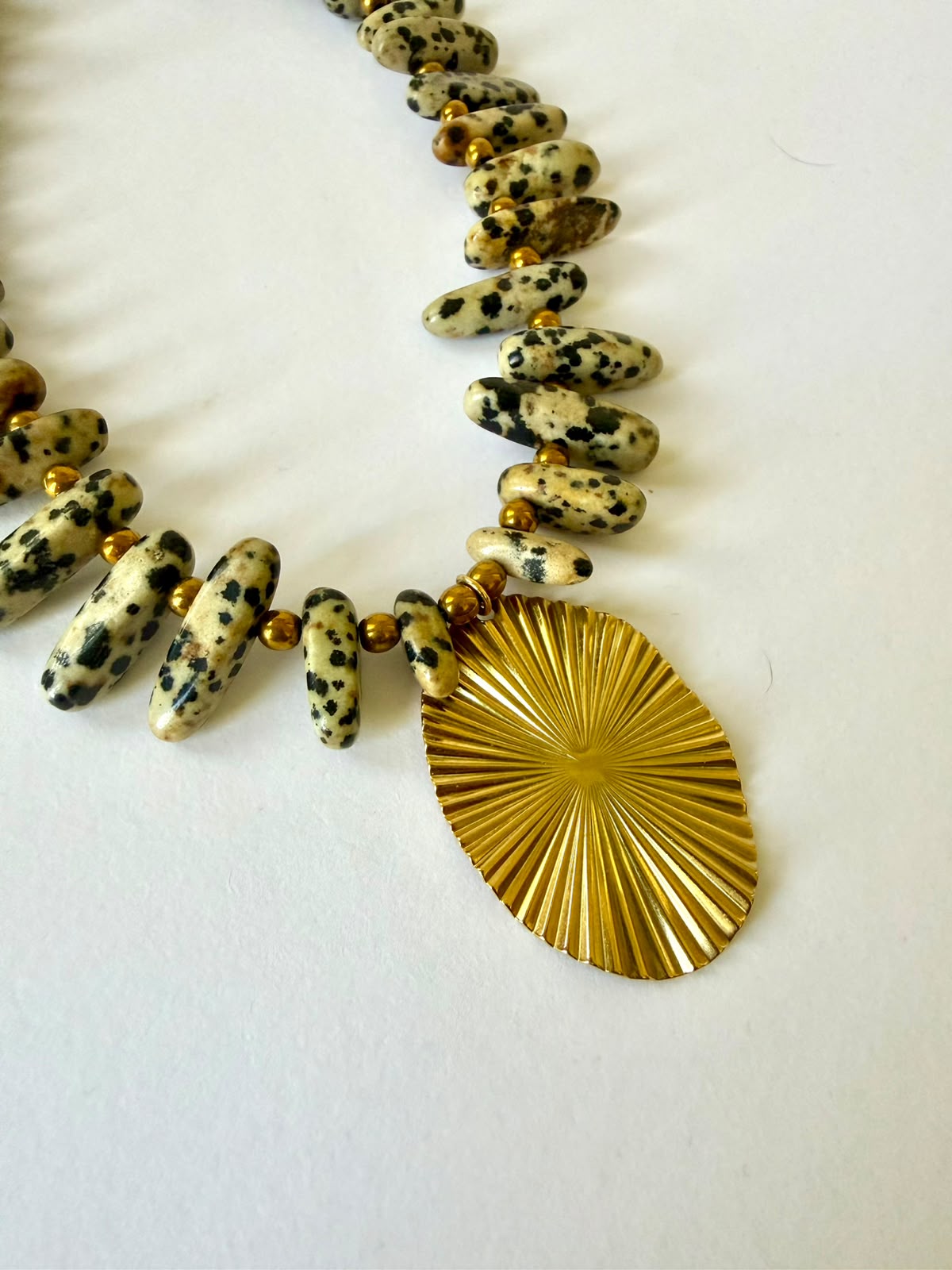 Close-up of elongated Dalmatian jasper beads, gold-tone spacers, and the oval sunburst pendant on a white background.