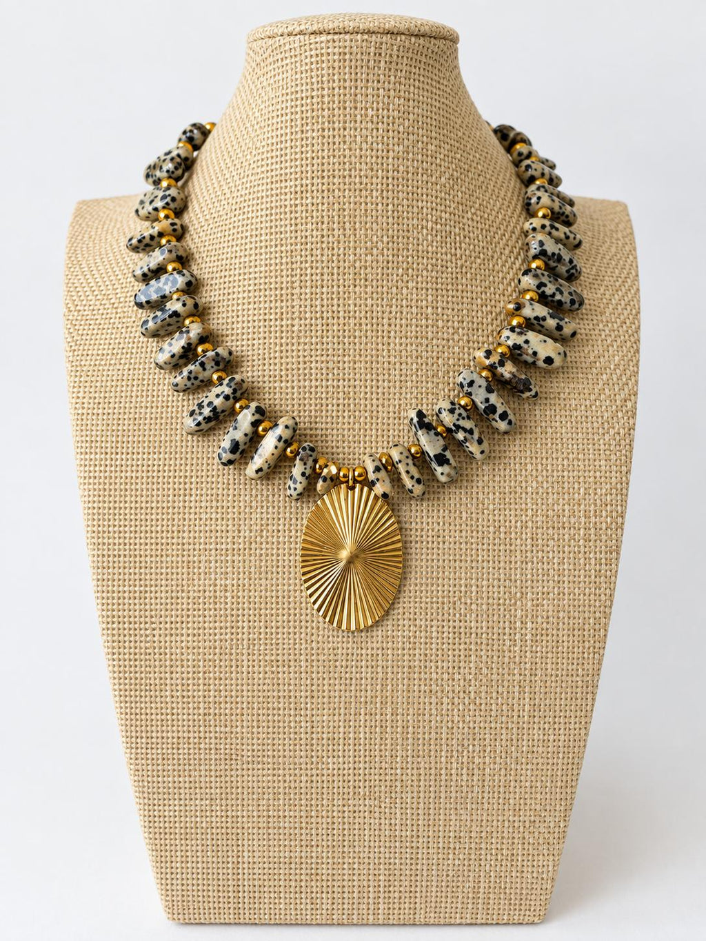 Necklace with elongated beige and black spotted beads, small gold-tone spacers, and an oval gold-tone sunburst pendant.
