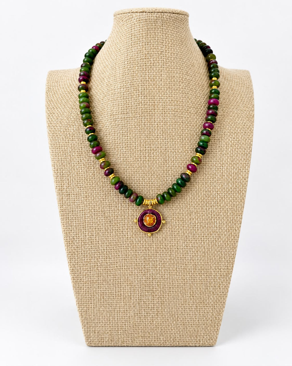 Green and purple jade beaded necklace with gold-tone accents and a round purple pendant with an orange center on a beige jewelry bust.