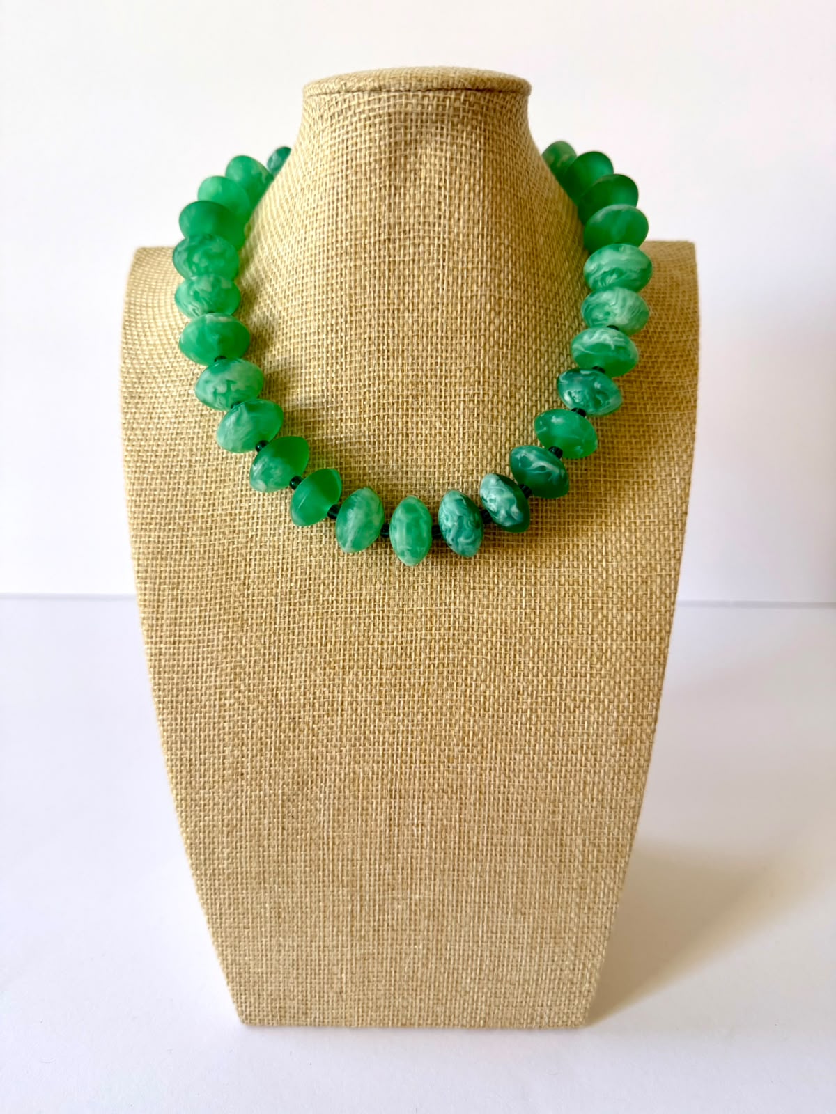 Green acrylic beaded necklace with rounded oval beads and pale marbled swirls on a beige jewelry bust.