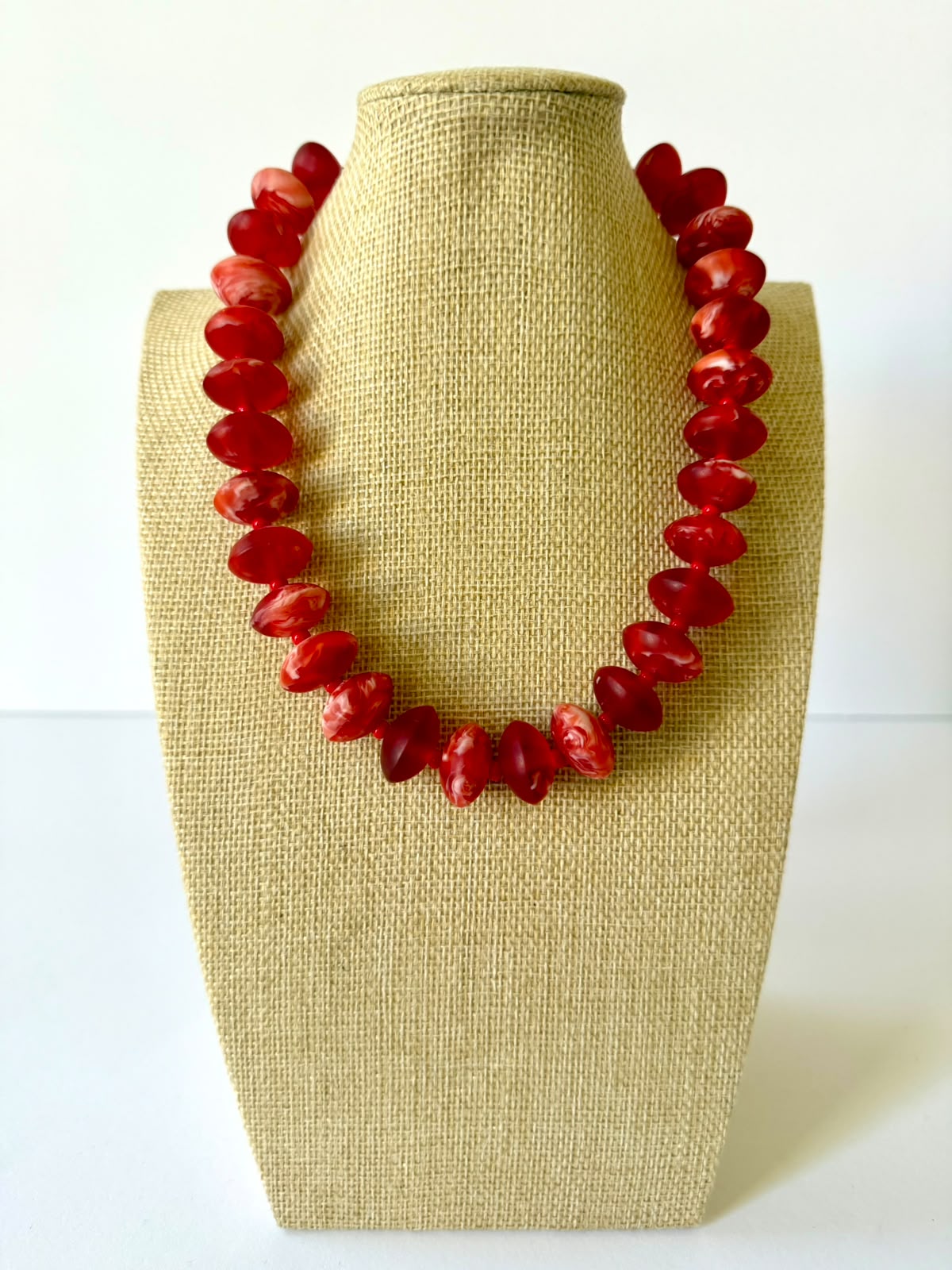 Red acrylic beaded necklace with rounded oval beads and pale marbled swirls on a beige jewelry bust.