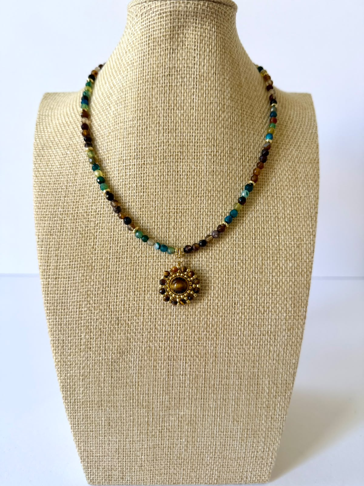 Multicolor faceted crystal beaded necklace with a round gold-tone pendant featuring a simulated tiger's eye focal stone on a beige jewelry bust.