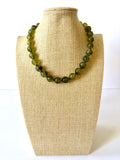 Olive green acrylic beaded necklace with rounded marbled beads and small gold-tone spacers on a beige jewelry bust.